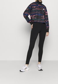 Žena v černých legínách, bílých teniskách a barevném černém mikině s červenými a modrými vzory, stojící s jednou rukou na boku.