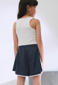 Witte geribde tanktop en marineblauwe shorts met elastische tailleband en zijaccenten. De shorts hebben een geplooid ontwerp en een lichtgrijze rand.