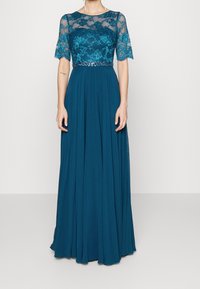 Femme portant une robe longue couleur sarcelle avec un haut en dentelle florale, manches courtes, et une ceinture à sequins, debout devant un fond uni.