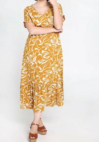 Musztardowa sukienka w białe kwiaty, z dekoltem w serek, krótkimi rękawami i warstwowym falbanym dołem. Modelka ma na sobie brązowe sandały na platformie.