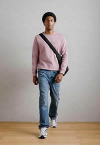 Gaiši rozā džemperis, zili džinsi un balti sporta apavi. Melna plecu soma un neitrā fona. Cilvēks stāv un iet uz priekšu.