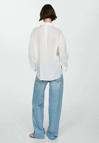 Camicia lunga bianca e trasparente con colletto, abbinata a jeans a gamba larga blu chiaro. L'outfit presenta una vestibilità rilassata e un design semplice.
