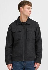 Chaqueta negra con cremallera hecha de una tela lisa y texturizada, que presenta dos bolsillos frontales y un cuello puntiagudo, llevada sobre una camiseta negra.
