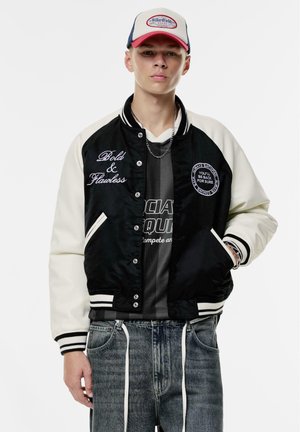 Veste varsity noire et crème avec texte brodé, boutons à pression, poignets rayés, et un t-shirt graphique noir en dessous, associé à un jean gris.