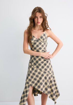 Jeune femme aux longs cheveux ondulés portant une robe asymétrique sans manches beige et noire à carreaux, debout les bras croisés devant un fond blanc.