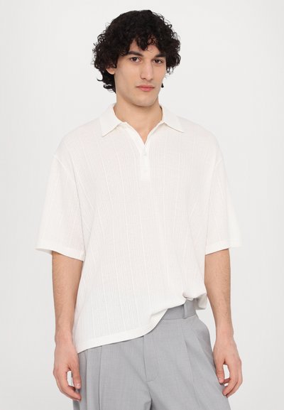 Polo shirt blanc à manches courtes avec un motif texturé et une patte de boutonnage à trois boutons, associé à un pantalon gris clair. Tissu léger.