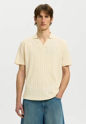 Junger Mann mit kurzen braunen Haaren, der ein cremefarbenes kurzärmeliges Strick-Poloshirt und blaue Jeans trägt, steht mit einer Hand in der Tasche vor weißem Hintergrund.