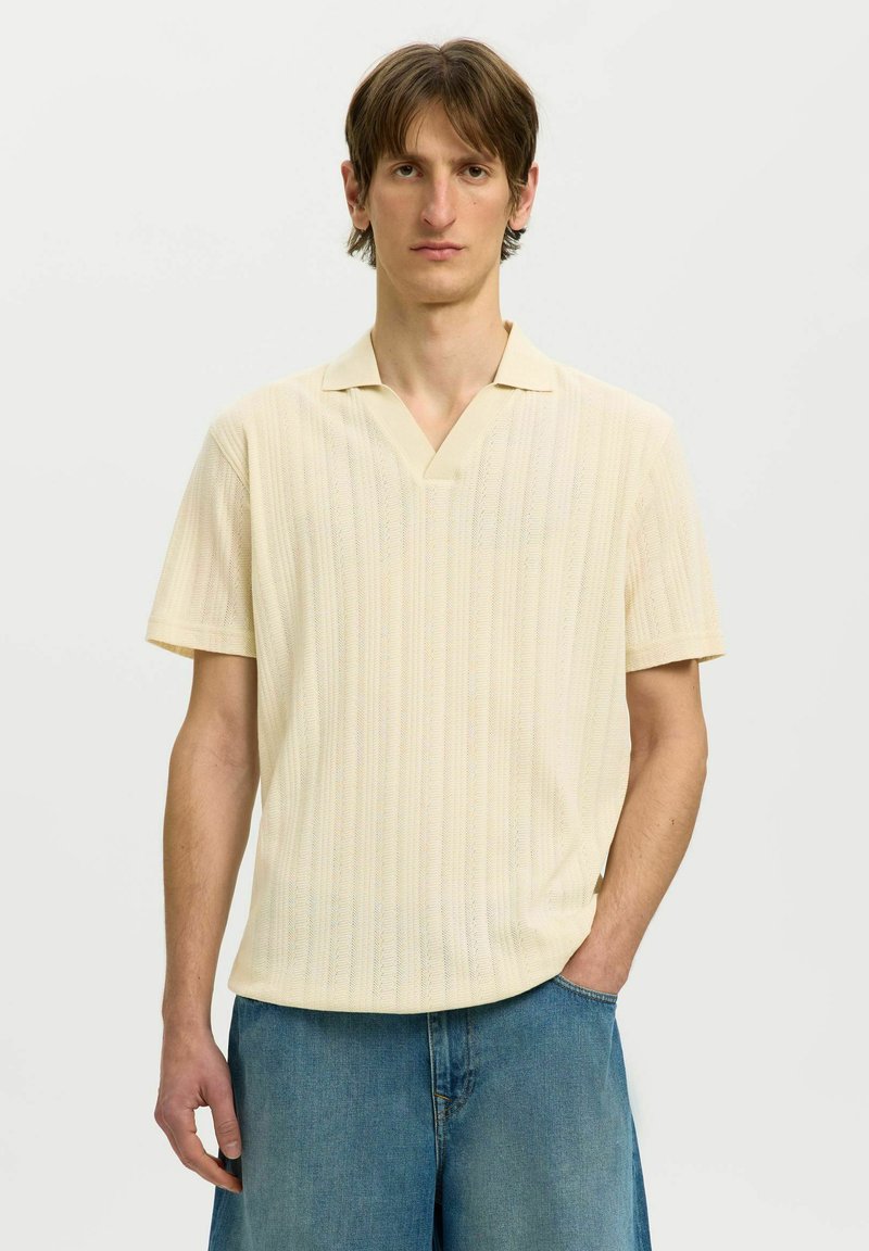 Jeune homme aux cheveux bruns courts, portant un polo en maille crème à manches courtes et un jean bleu, debout avec une main dans la poche sur fond blanc.