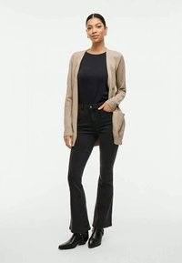Cardigã de malha bege com frente aberta, mangas longas e bolsos, usado sobre uma blusa preta e combinado com jeans pretos à boca de sino e botas de tornozelo.