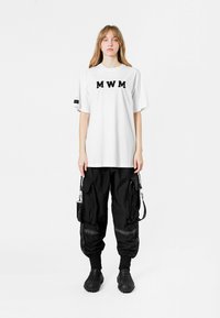 Camiseta blanca de algodón con letras negras "MWM", combinada con pantalones cargo negros con bolsillos con cremallera y puños elásticos. Calzado negro.