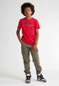 Rode T-shirt met korte mouwen bedrukt met de tekst "PETROL INDUSTRIES & CO", gecombineerd met olijfgroene cargobroek en zwarte sneakers met witte accenten.