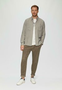 Lichtgrijs overhemd over een wit T-shirt, gecombineerd met olijfgroene ribfluwelen broek en witte sneakers. Glad fabric met een casual pasvorm.