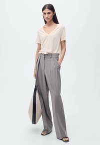 Haut beige à col en V et à manches courtes associé à un pantalon large gris taille haute. Le modèle tient un grand sac noir et beige. Textures lisses.