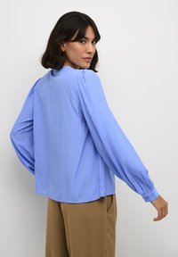 Blusa blu chiaro con maniche lunghe e gonfie; texture morbida; sottili righe verticali; scollatura rotonda; polsini con bottoni; vestibilità comoda.