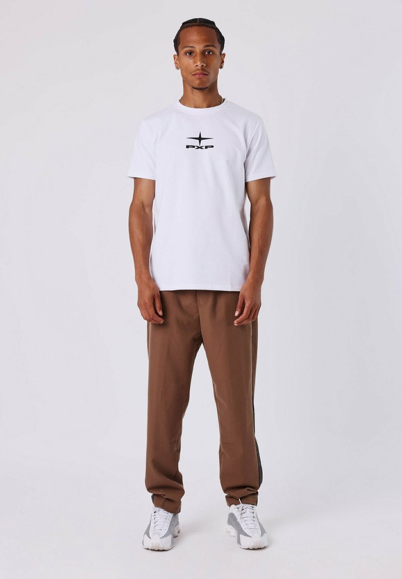 Witte t-shirt met korte mouwen en zwart "PXP" logo op de borst, gecombineerd met bruine tapered broek en grijze sportschoenen.