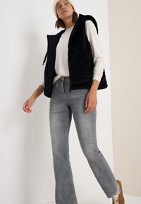 Faux fur marinblå väst med huva, zipförslutning, lagd över en ljusfärgad topp. Bärs med raka grå jeans och bruna sneakers.