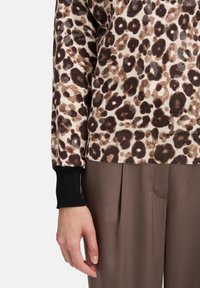 Sweter w panterkę w odcieniach beżu, brązu i czerni z prążkowanymi mankietami; w połączeniu z gładkimi, brązowymi spodniami plisowanymi, widoczna dłoń.