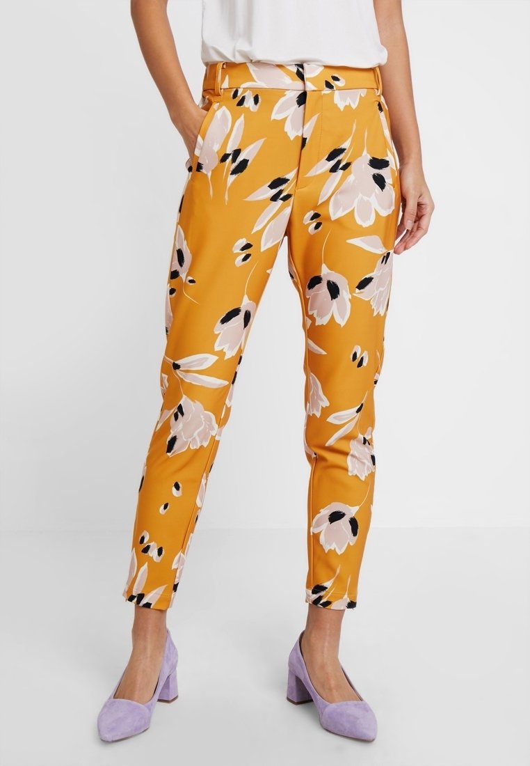 Pantalon floral jaune avec motifs roses et noirs, coupe slim, poches latérales et texture lisse, assorti à des talons lavande.