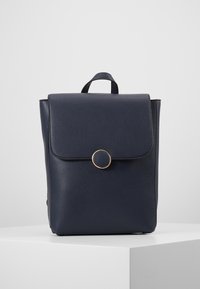 Marinblå läderryggsäck med en texturerad yta, med en framflik, cirkulär guldfärgad hårdvara och ett handtag för bärande.