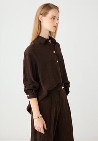 Brown collared blouse with long sleeves, textured fabric, gold button accents, and matching high-waisted trousers, complemented by a gold bracelet.