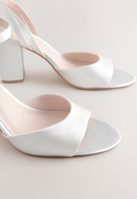 Hvide satin åbne peep-toe sandaletter med blokhæl og ankelrem på en lys neutral baggrund.