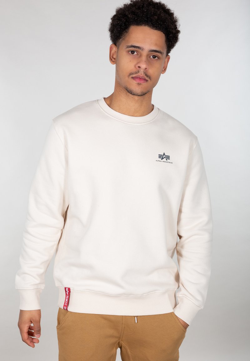 Cremefarvet sweatshirt med rund halsudskæring, ribbede manchetter og et lille sort logo på brystet. Har et rødt mærke ved kanten.