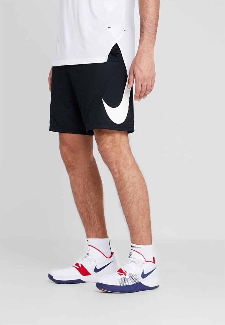 Homme portant un short noir Nike avec un swoosh blanc, des chaussures de sport blanches avec des accents bleus et rouges, et des chaussettes blanches, se tenant sur un sol gris.