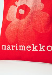 Piros vászontáska, amelyen egy nagy fehér virágmintázat található, az alján pedig vastag, törtfehér betűkkel a "marimekko" felirat olvasható.