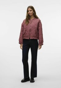 Giacca imbottita in malva con motivo a rombi, chiusura con zip e tasche laterali; abbinata a jeans svasati scuri e scarpe nere.