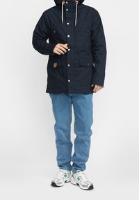 Mörkblå parka-jacka med huva, med knäppningar, två framfickor och en beige detalj. Matchad med ljusblå jeans och sneakers.
