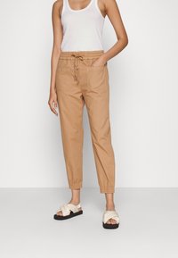 Lightweight tan pants with elastic waistband, drawstring detail, and two front pockets. Paired with white cross-strap sandals.