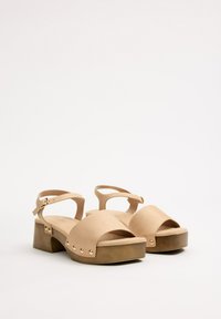 Las sandalias de plataforma beige presentan una amplia correa frontal, una correa de tobillo con hebilla y acentos de tachuelas doradas en una base texturada que imita la madera.