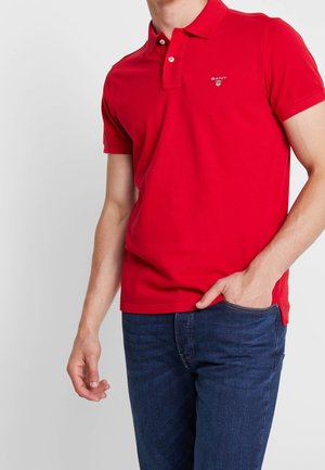 Homme portant un polo rouge à manches courtes avec un petit logo sur la poitrine, associé à un jean bleu foncé, une main dans la poche.
