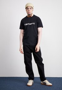 Czarny bawełniany t-shirt z białym logo "carhartt", zestawiony z czarnymi spodniami i białymi sneakerami z ciemnoniebieskimi akcentami. Teksturowana beżowa czapka.