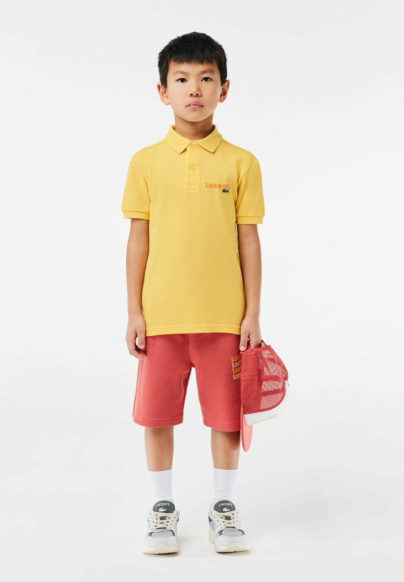 Polo shirt jaune, shorts roses, chaussettes blanches et baskets grises. Présente le logo de la marque sur la chemise, et une casquette rose à la main.