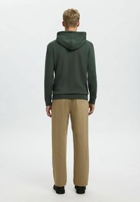 Felpa verde scuro con cappuccio regolabile, abbinata a pantaloni beige. L'outfit presenta una vestibilità rilassata e polsini elastici discreti.