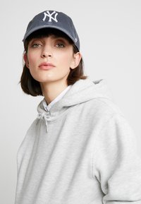 Sweatshirt à capuche gris avec cordon de serrage, en tissu texturé, associé à une casquette bleu foncé avec un logo brodé blanc.