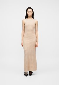 Γυναίκα στέκεται φορώντας αμάνικο μπεζ μάλλινο μάξι φόρεμα και μαύρα μυτερά παπούτσια με λουράκια και αγκράφες σε απλό λευκό φόντο.