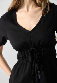 Black v-neck top with short sleeves, cinched waist featuring a drawstring detail, made from soft stretch fabric for a comfortable fit.