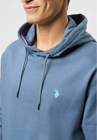 Kapuca v teal tkanini, ki ima sprednji kenguru žep, vrvico za zategovanje in majhen vezen logotip na prsih.