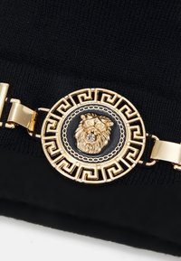 Curea neagră cu un emblema a unui leu în ton de aur. Emblema prezintă detalii complicate într-un design circular, încadrat de un model în formă de cheie grecească.
