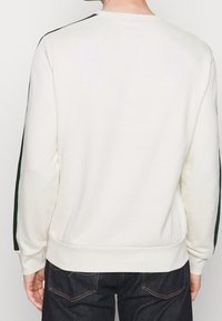 Bílá mikina s kulatým výstřihem s zelenými bočními pruhy, žebrovanými manžetami a lemem a hladkým povrchem, nošená s tmavými džínami.