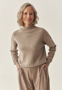 Beige, bordázott pulóver magas nyakkal, chevron mintával és laza szabással, taupe színű, pliszézett nadrággal párosítva.