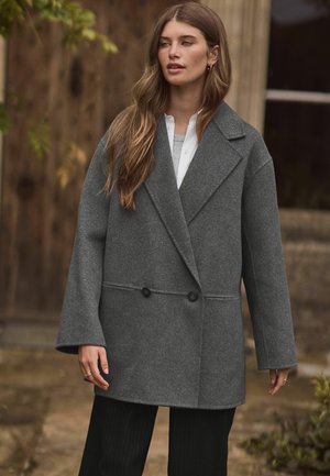 Grijze dubbelgeknopte wollen jas met brede revers, oversized mouwen en twee knopen. Gecombineerd met een witte blouse en zwarte streepjesbroek.