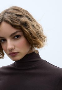 Brązowy top z wysokim kołnierzem wykonany z gładkiej tkaniny, o dopasowanym kroju i minimalistycznym designie, uzupełniony naturalnymi, falującymi włosami.