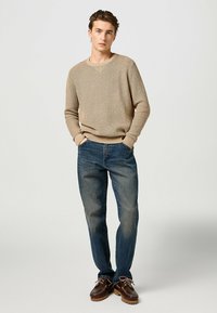 Beige gestrickter Pullover mit einem strukturierten Muster, kombiniert mit dunkelblauen Jeans. Das Modell trägt braune Bootsschuhe mit Lederakzenten.