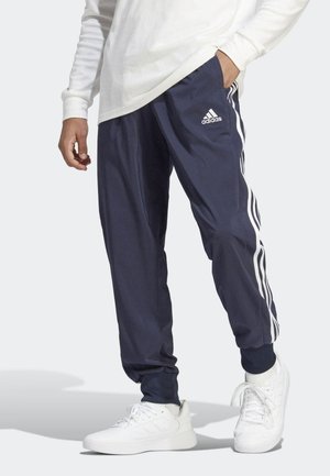 Osoba ubrana w granatowe spodnie dresowe Adidas z białymi paskami i białe sneakersy, stojąca z jedną ręką w kieszeni.
