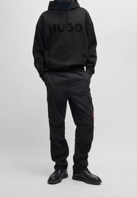 Sudadera con capucha negra con el logotipo "HUGO", combinada con pantalones cargo negros que tienen bolsillos laterales. El calzado consiste en botas de cuero negras.