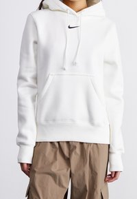 Persona vestida con un hoodie blanco de Nike con bolsillo delantero y pantalones cargo beige, de pie contra un fondo claro y sencillo.