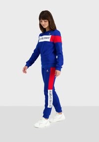 Tuta blu in cotone, con dettagli rossi e branding bianco. La parte superiore ha un collo tondo; i pantaloni hanno polsini elastici.